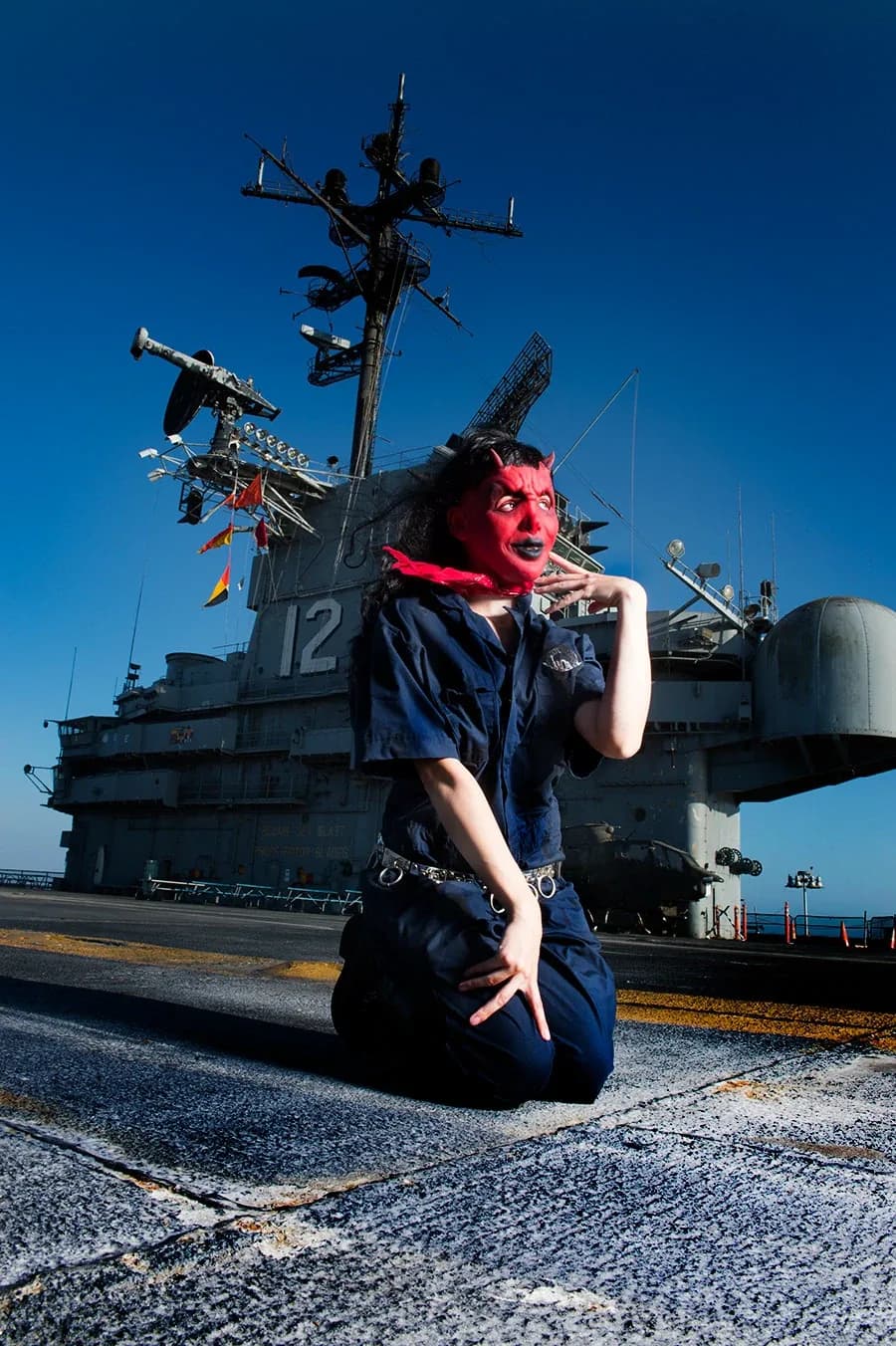 A woman in red devil makeup kneeling on the flight deck, the carrier's island towering behind her.
