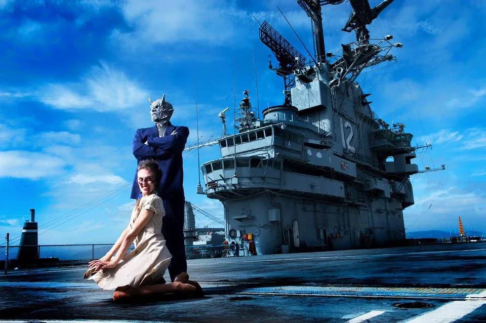 A uniformed sailor standing over a woman kneeling on the flight deck, the ship stretching out behind them.